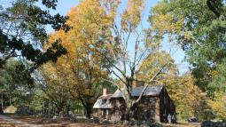 Two story, unpainted wooden house beside a dirt road bordered by stone walls. Trees and fall colors