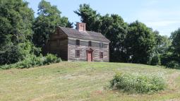 A two story wooden colonial house sits on a hill surrounded by green trees.