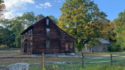 A wooden colonial era house stands in a grassy field surrounded by trees and a wooden rail fence