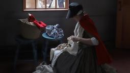 A women in 1775 clothing sits near a window in the Smith House wrapping bread with a cloth