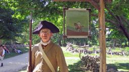 Revolutionary War soldier with a musket standing beneath a painted wooden tavern along a dirt roada