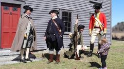 Three Militia and one British reenactor stand in front of a wooden house speaking to a child