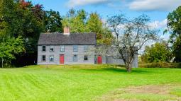 A wooden colonial era salt box house with red painted door sits in a green field of grass