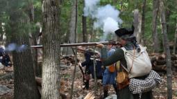 A militia soldier leans against tree and fires a musket
