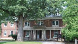A large brick mansion house with covered porch and white pillars. A large tree stands at the front