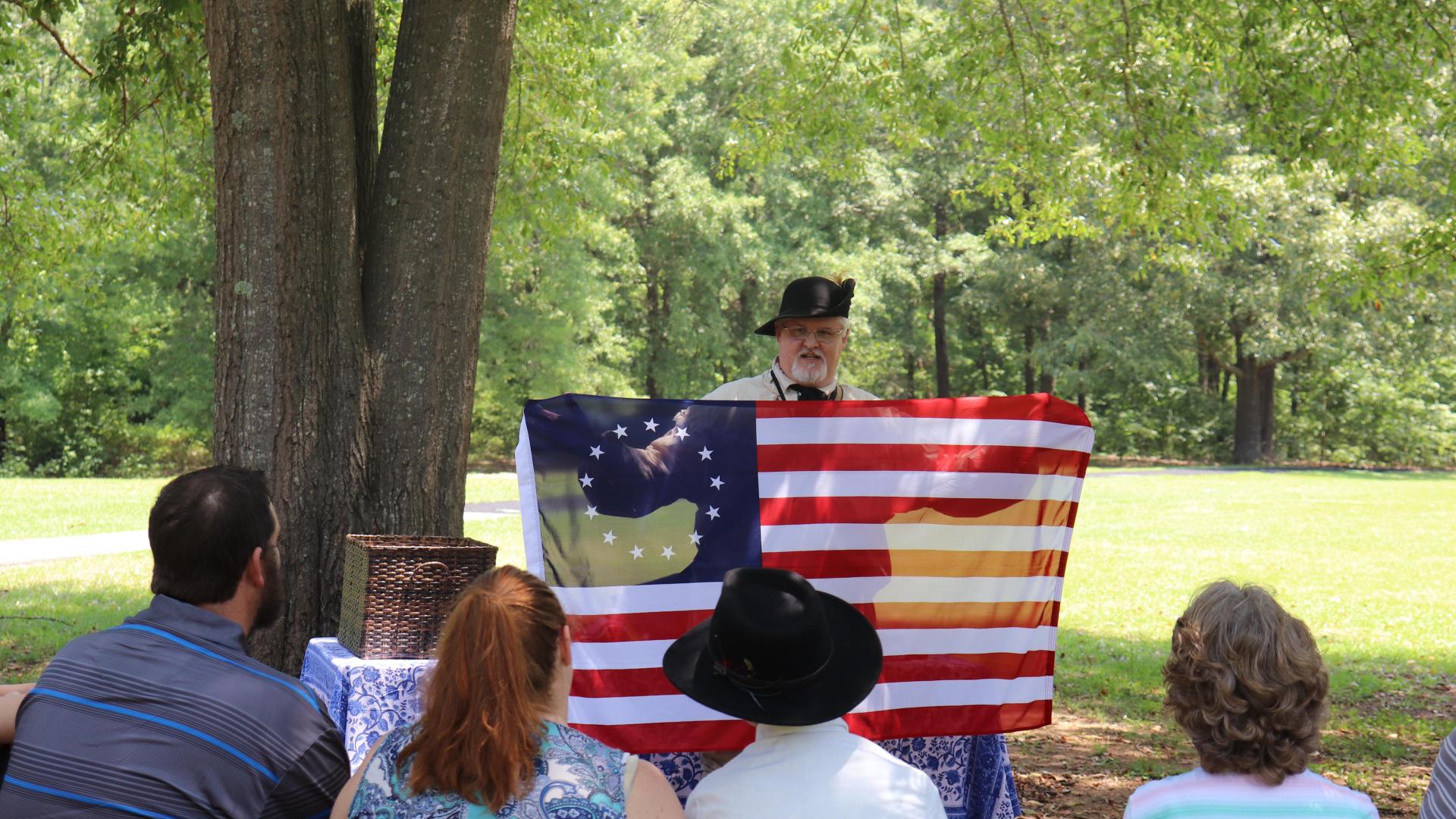Volunteer dressed as a 18th century militia person holds a flag for a group of visitors.
